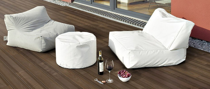 Dunkle Holzterrasse neben einem Gebäude mit zwei Sitzkissen und dekorativen Elementen.