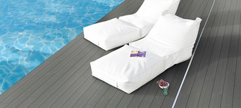 Elegante Poolterrasse mit dunklem WPC-Belag, zwei weißen Lounge-Liegen und ruhiger Wasserreflexion.