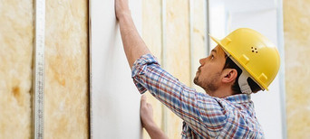 Handwerker mit gelbem Schutzhelm montiert eine Gipskartonplatte auf eine Metallständerwand mit Dämmung.