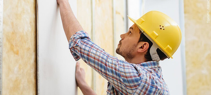 Handwerker mit gelbem Schutzhelm montiert eine Gipskartonplatte auf eine Metallständerwand mit Dämmung.