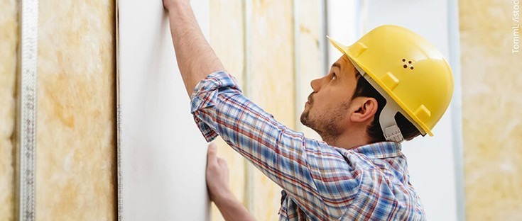 Handwerker mit gelbem Schutzhelm montiert eine Gipskartonplatte auf eine Metallständerwand mit Dämmung.